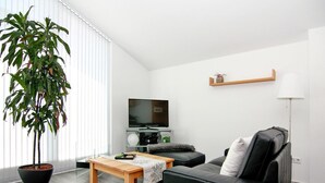 A modern living room with a black leather sofa, a wooden coffee table, a large potted plant, a flat-screen TV, and a floor lamp. - Appartementhaus Calmsailing, Borgerende (Boergerende-Rethwisch)