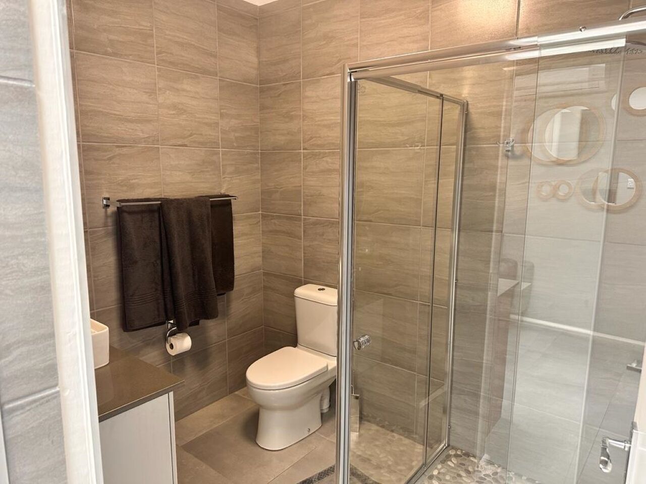 Un baño moderno con ducha envuelta en vidrio, inodoro y lavamanos con toallero.