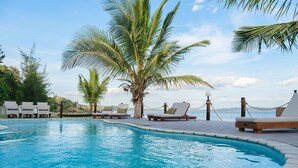 Zona junto a la piscina con sillas de descanso, una palmera y vista al mar.