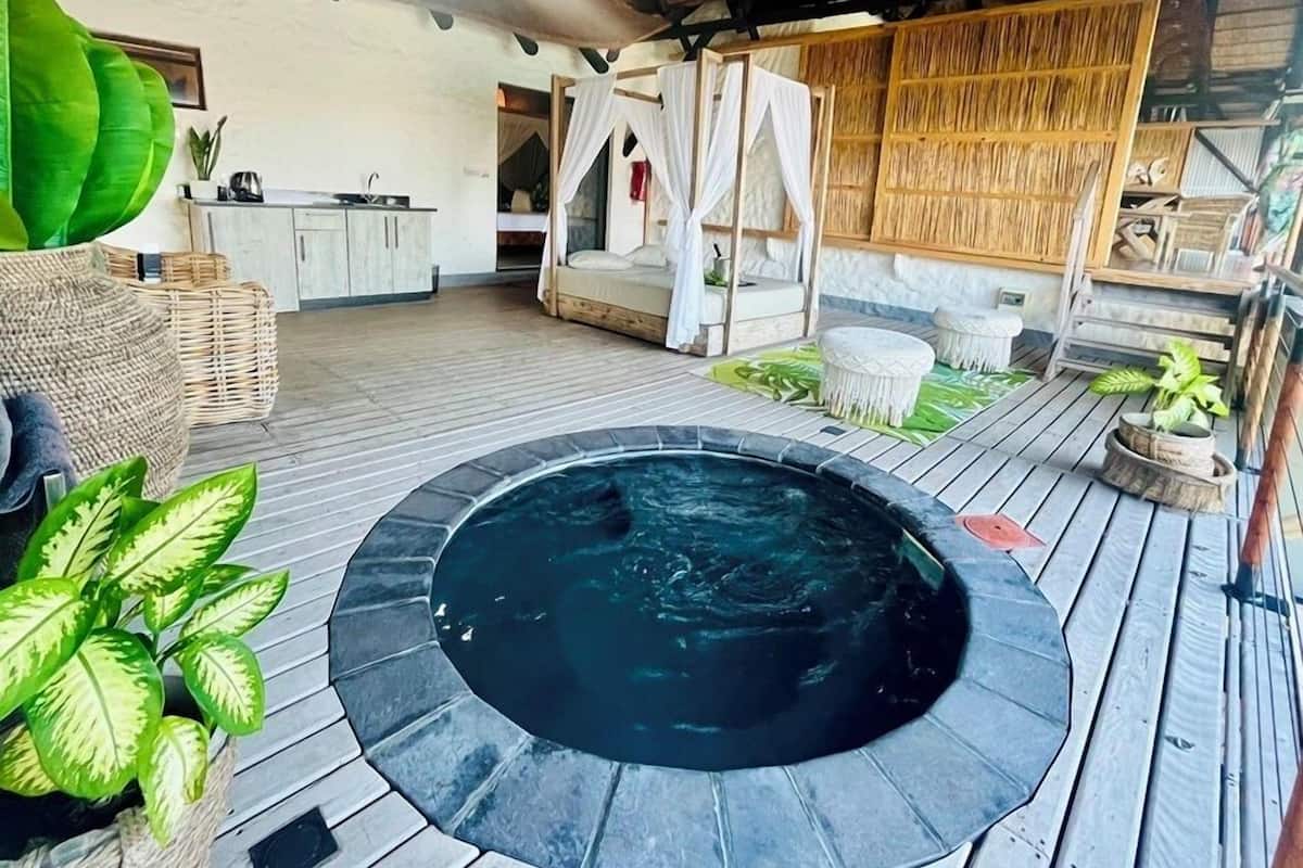 Una bañera de hidromasaje rodeada de una terraza de madera, una cama con dosel y una zona de cocina rústica.