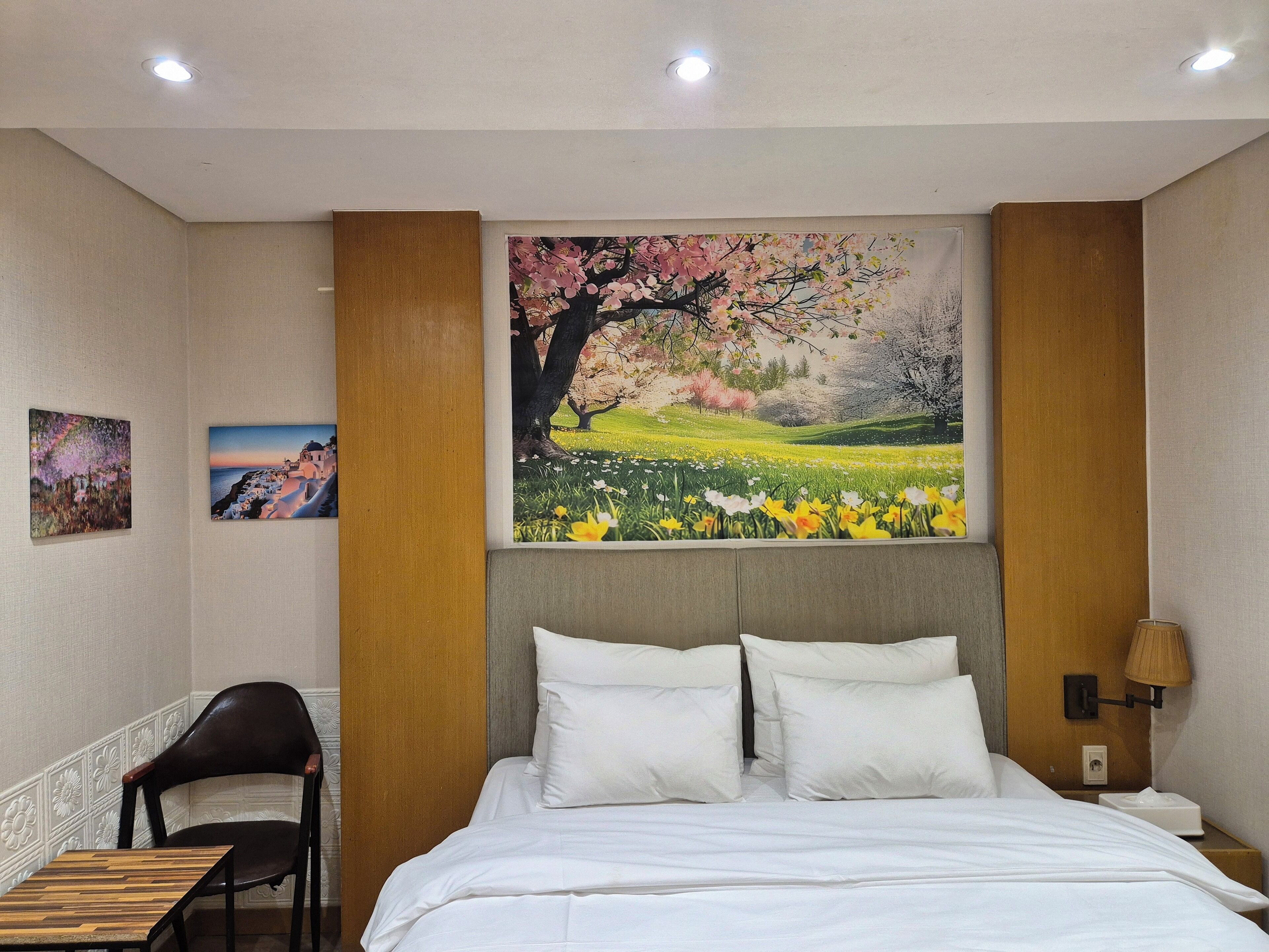 Una habitación de hotel con una cama, una mesita de noche de madera, una lámpara y dos cuadros en la pared.