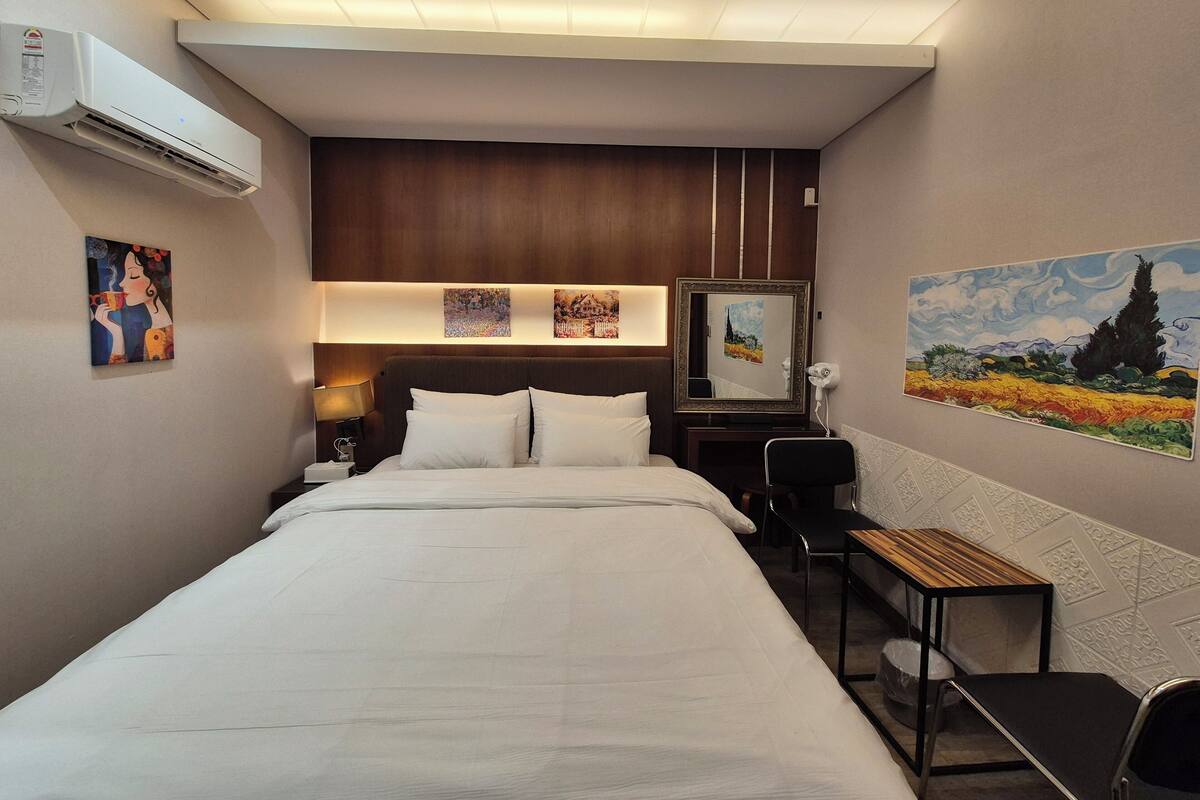 A hotel room with a large bed, a desk, and a chair. There are paintings on the walls and an air conditioning unit mounted on the wall.