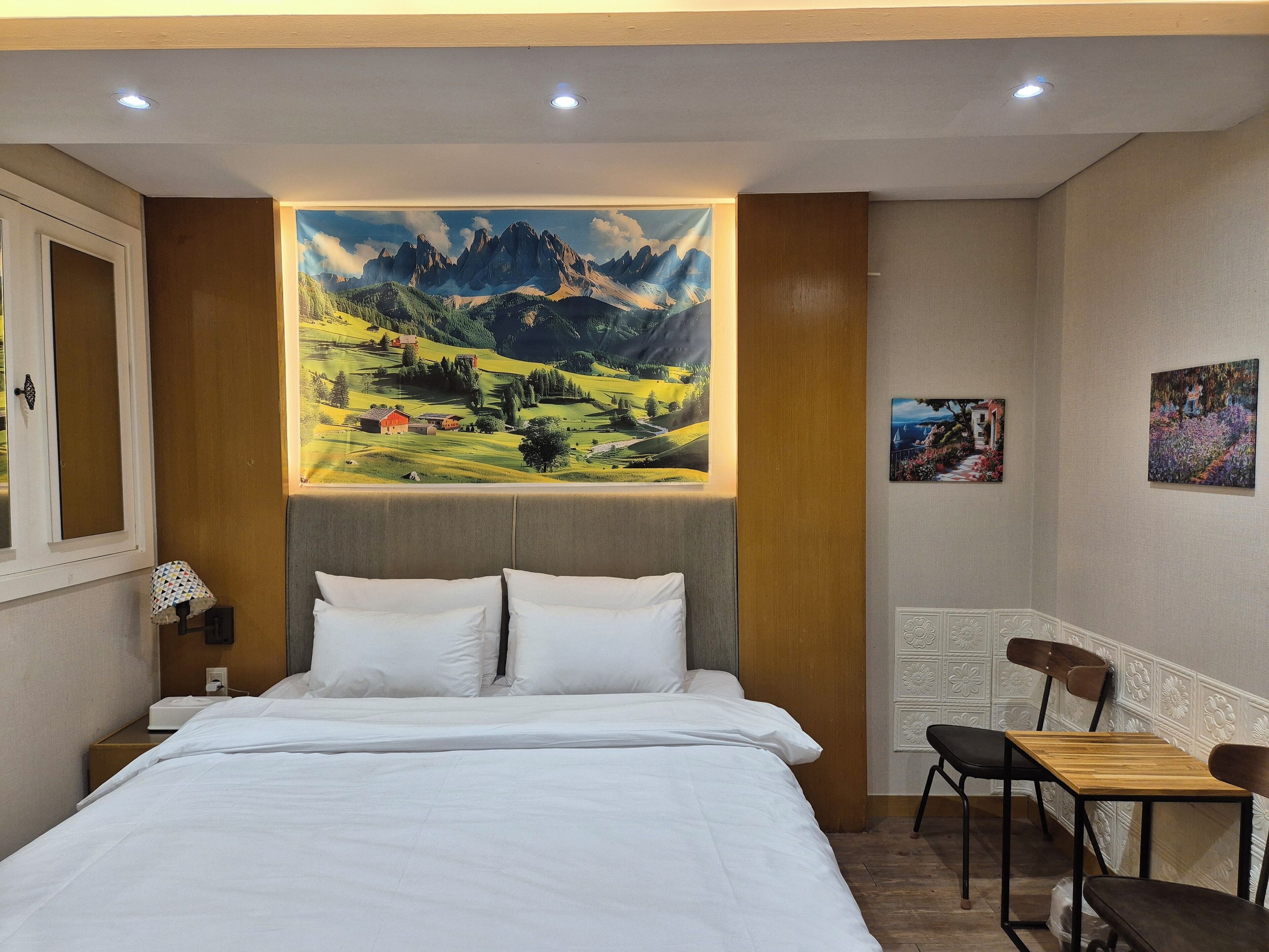 A hotel room with a bed, a desk, and chairs. There is a large painting of a mountain landscape on the wall.