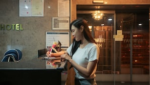 Un comptoir d’accueil d’hôtel avec une femme derrière, un panneau affichant le menu et une enseigne « HÔTEL » sur le mur.