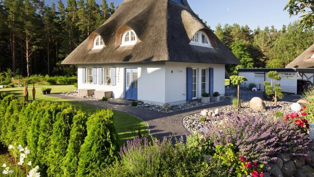 Una casa con techo de paja y fachada blanca, rodeada de un jardín cuidado con diversas plantas y flores.