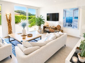 Una sala de estar moderna con un sofá, una mesa de centro y un televisor. Hay plantas en macetas y una vista de follaje verde al exterior.