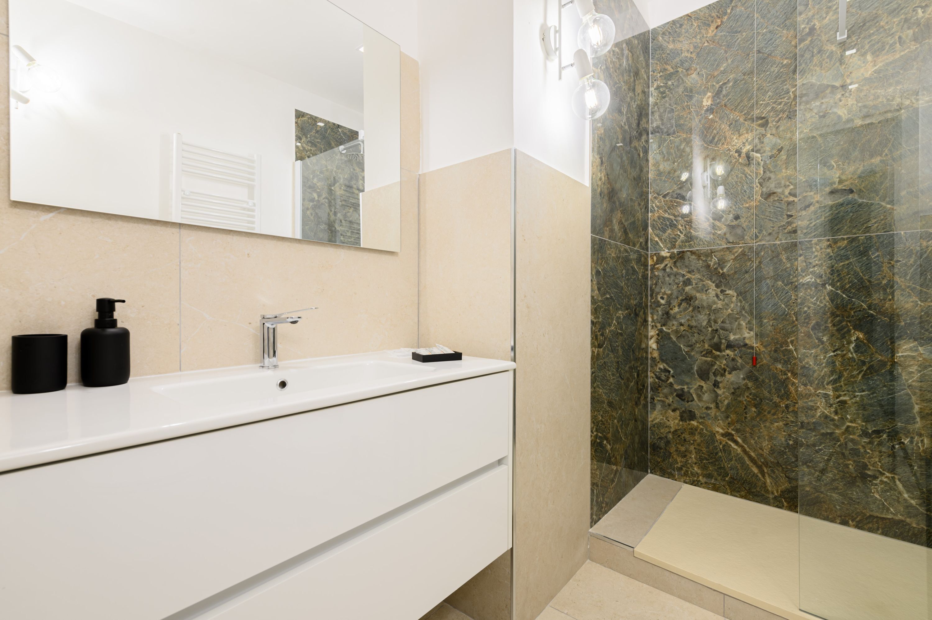 A modern bathroom with a white vanity, a mirror, a black soap dispenser, and a shower with a marble wall.