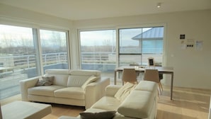 Ein geräumiges Wohnzimmer mit einer großen weißen Couch, einem kleineren weißen Sessel und einem Couchtisch. Es gibt einen Essbereich mit Tisch und Stühlen sowie große Fenster mit Blick ins Freie.