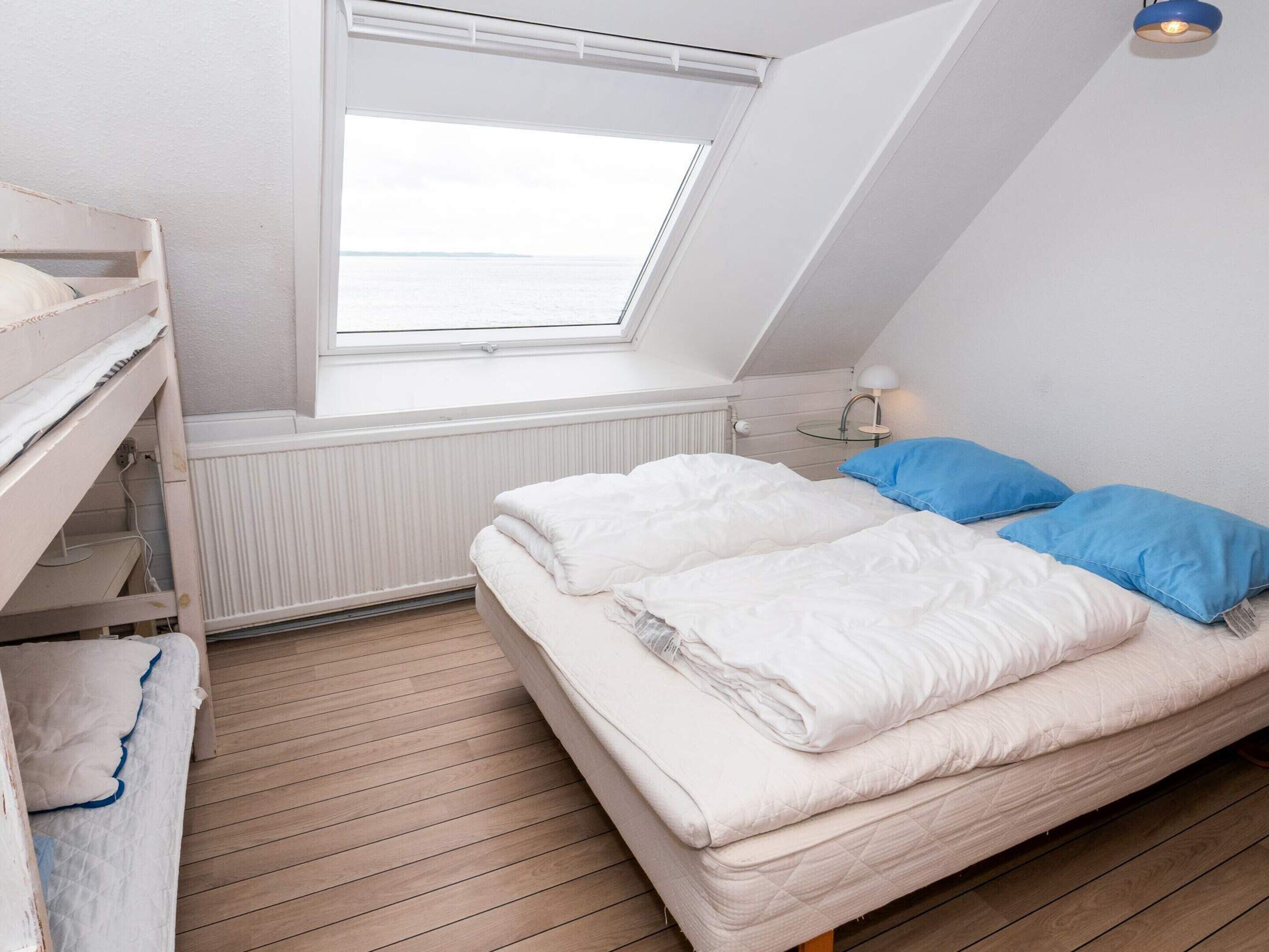 A bedroom with a bunk bed, a single bed with blue pillows, a skylight, a bedside lamp, and a view of the sea.