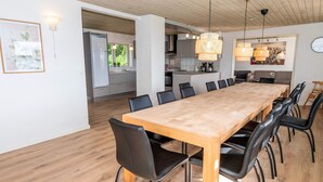 Uma ampla área de jantar com uma longa mesa de madeira e cadeiras pretas, uma cozinha ao fundo e um design de teto moderno.