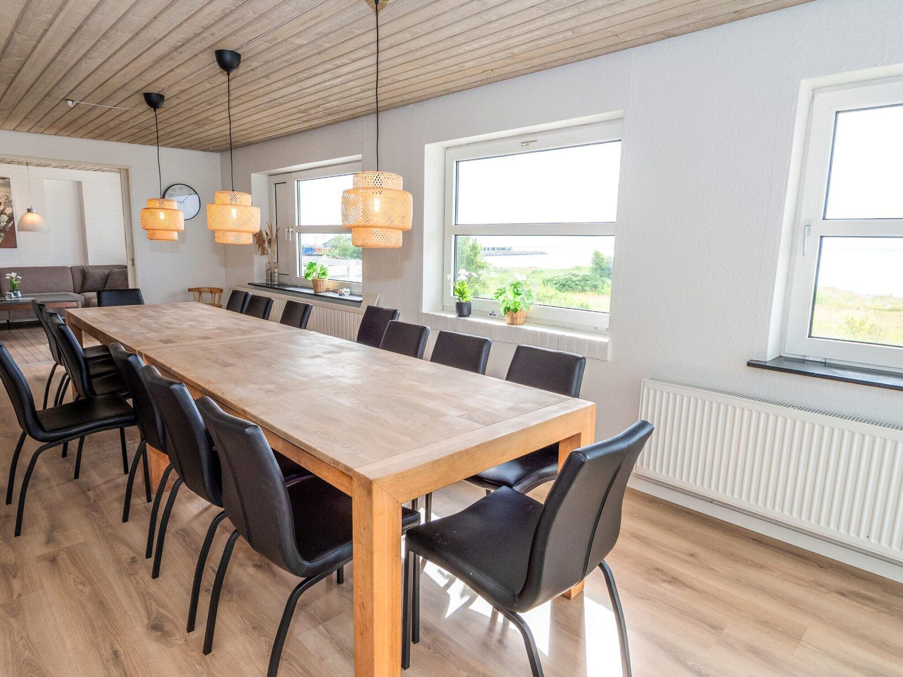 Uma sala de jantar ampla com uma longa mesa de madeira e cadeiras pretas, grandes janelas com vista e luminárias pendentes.