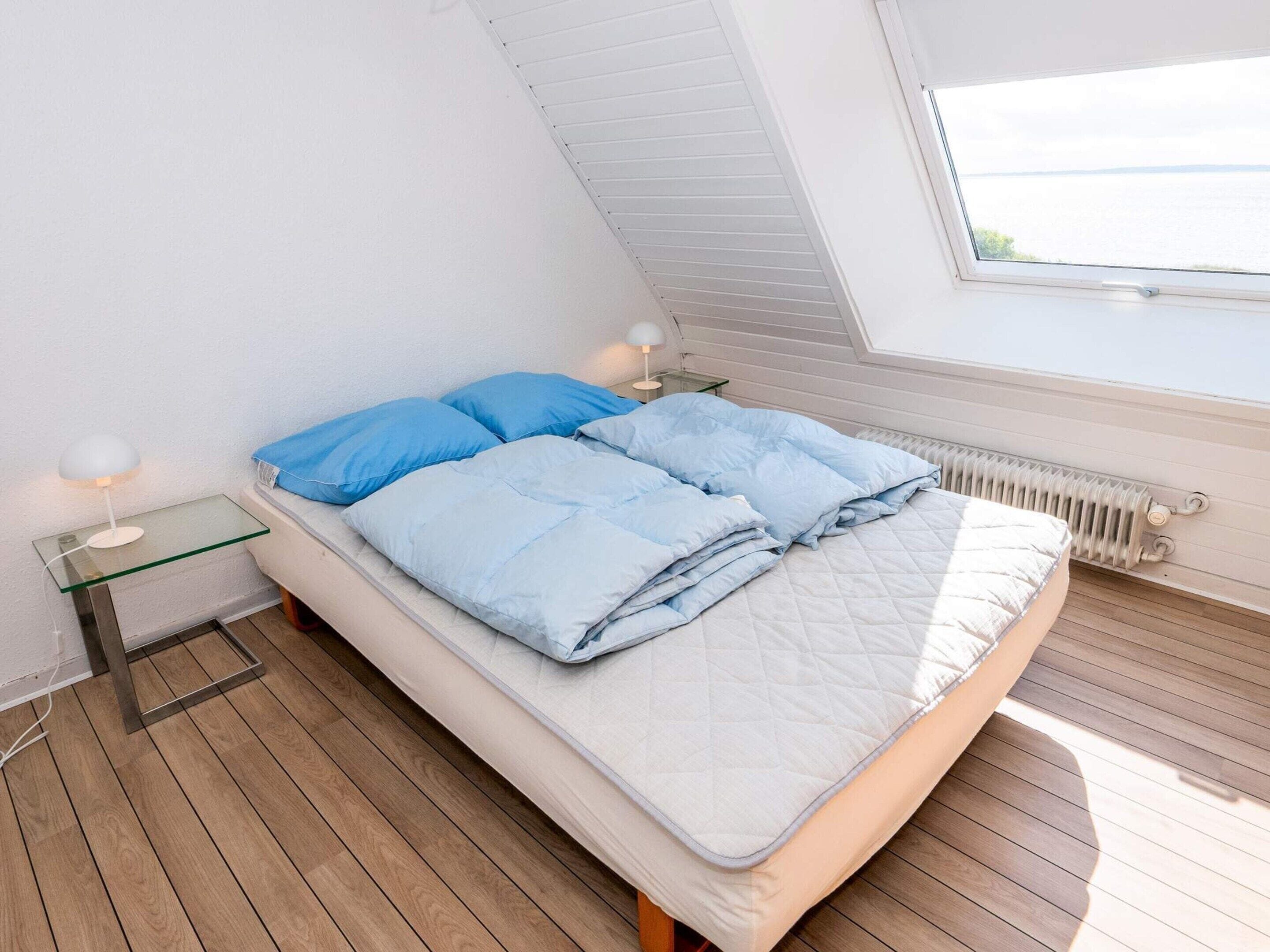 A bedroom with a sloped ceiling, a bed with blue bedding, a bedside table with a lamp, and a view of the sea through a window.