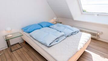 A bedroom with a sloped ceiling, a bed with blue bedding, a bedside table with a lamp, and a view of the sea through a window.
