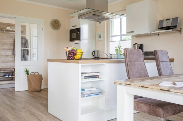 A modern kitchen with white cabinetry, a built-in microwave, and a coffee maker. There is a dining area with a table and chairs, and a small island with a basket of fruits and books.