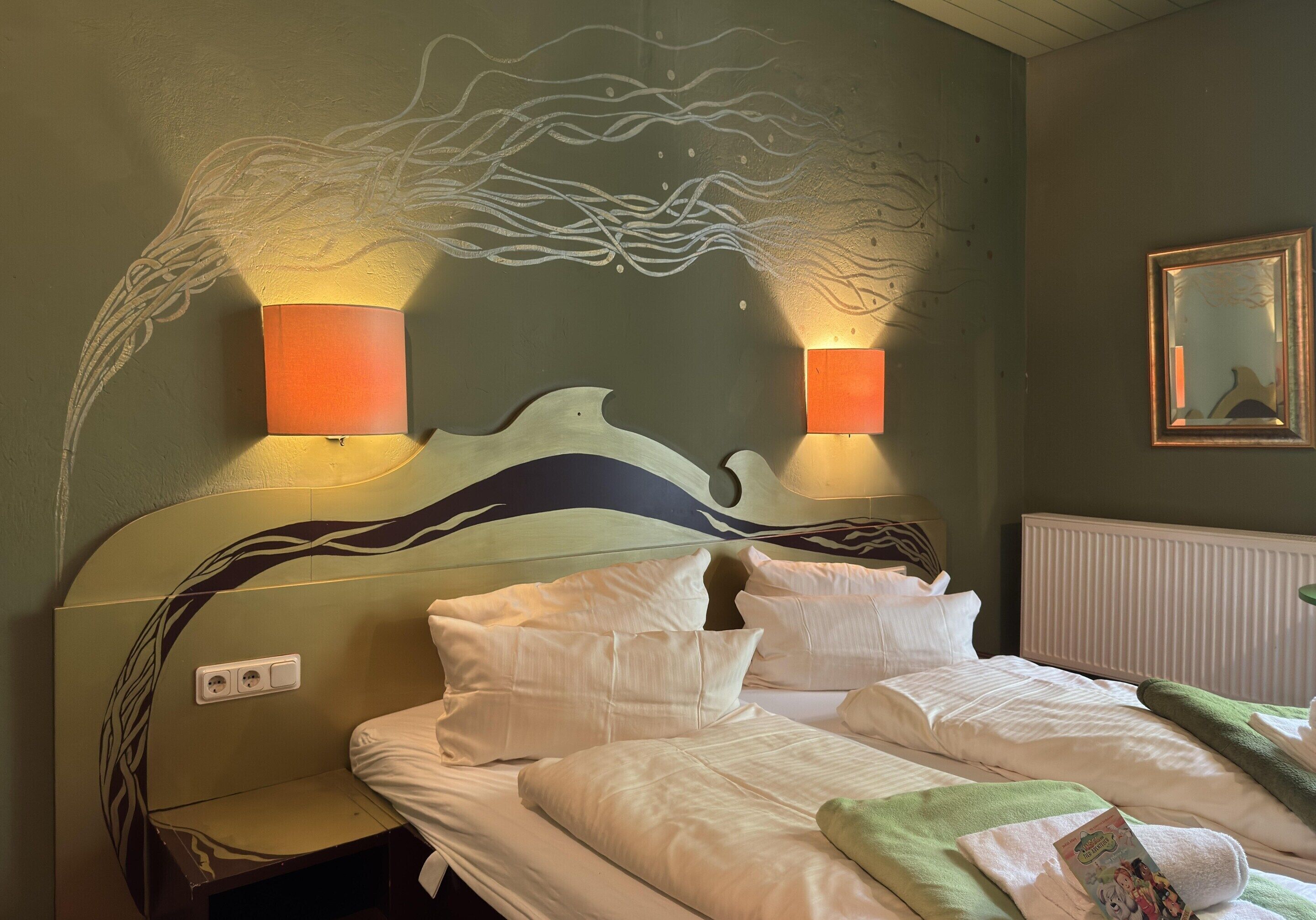 A hotel room with a bed, two bedside lamps, a wall mural, a radiator, and a framed picture on the wall.