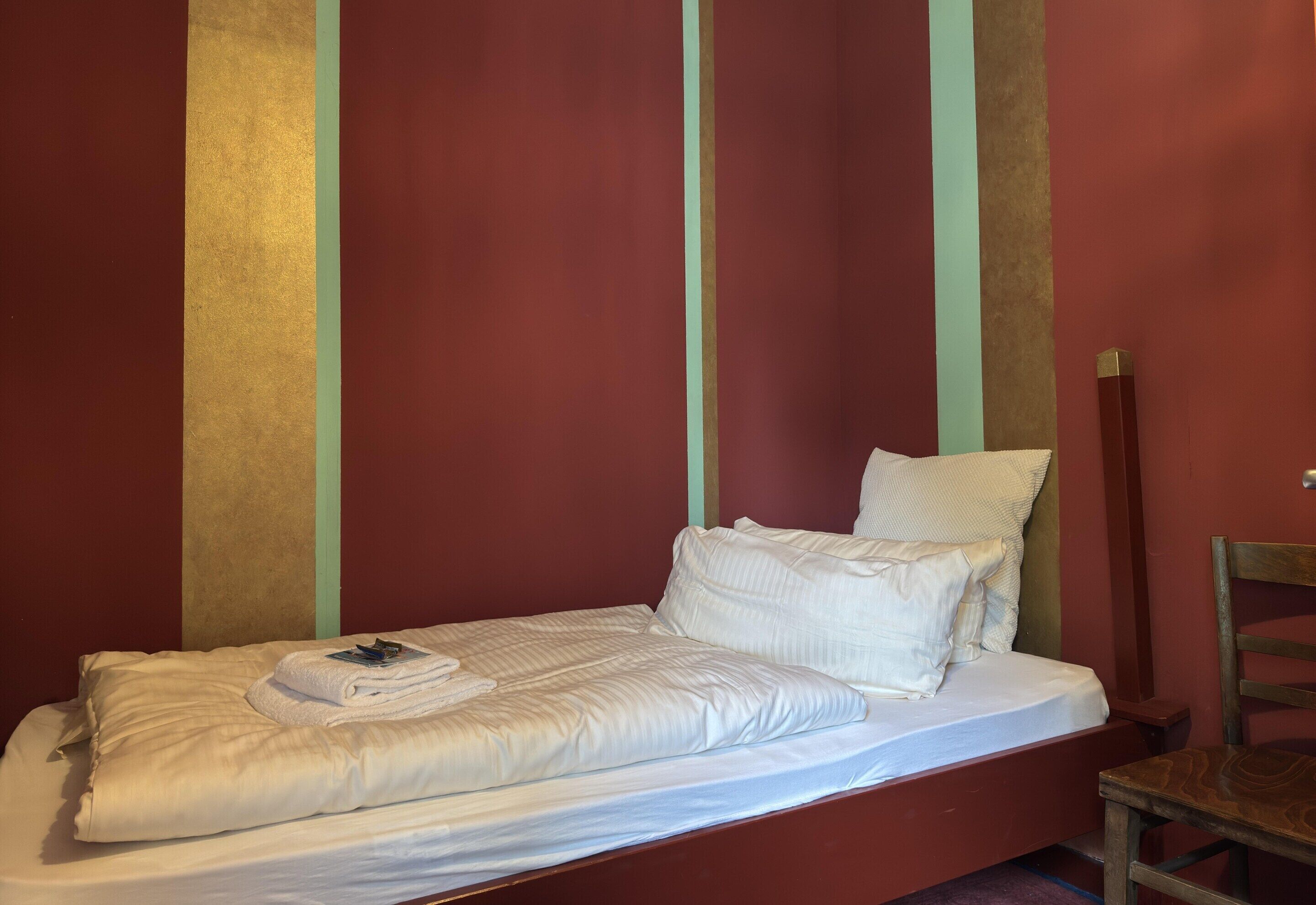 Uma cama de solteiro com roupa de cama branca num quarto com paredes vermelhas e uma cadeira de madeira.