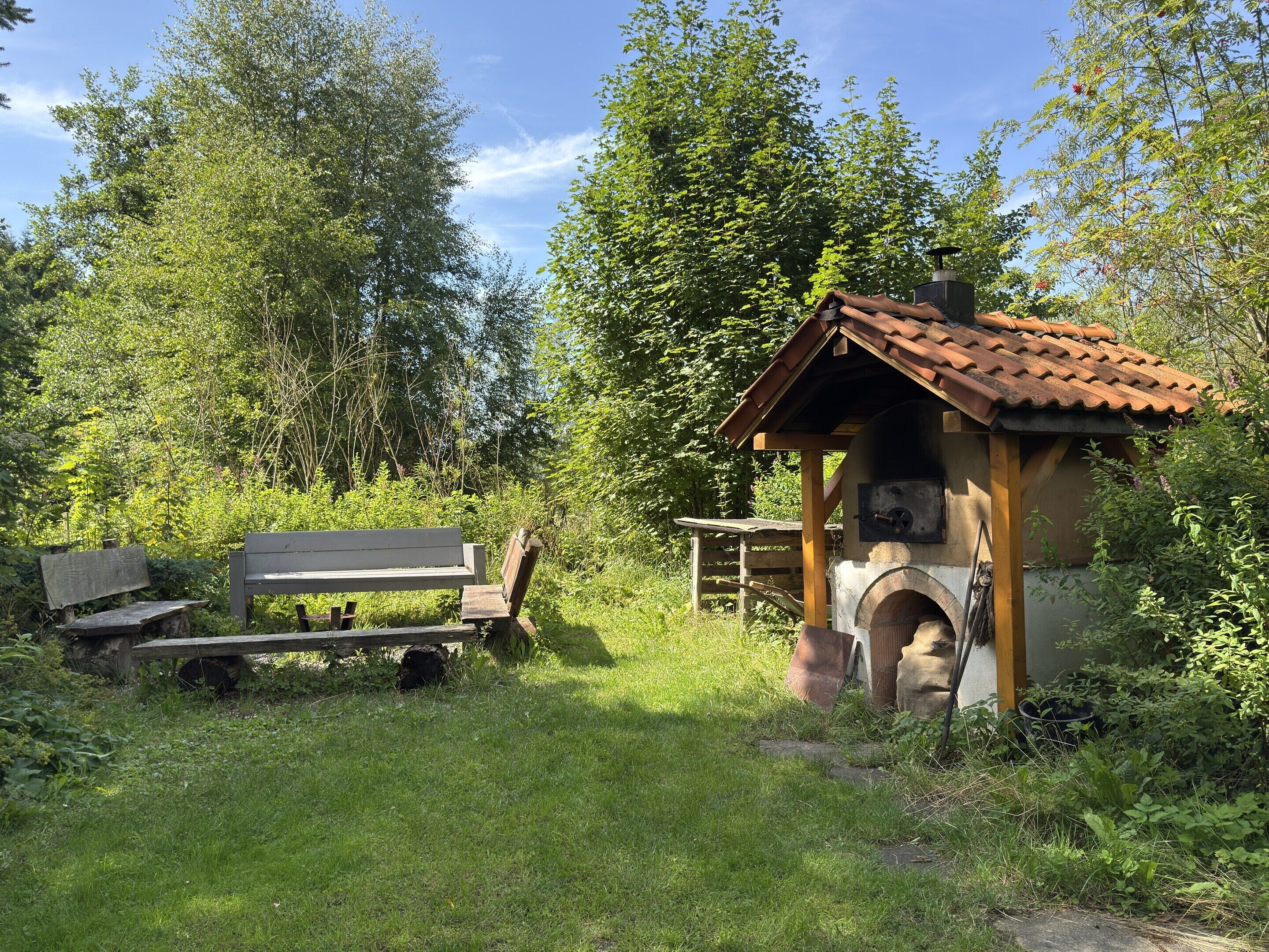 A rustic outdoor seating area with a wooden bench and a small wooden structure with a tiled roof, surrounded by lush greenery.