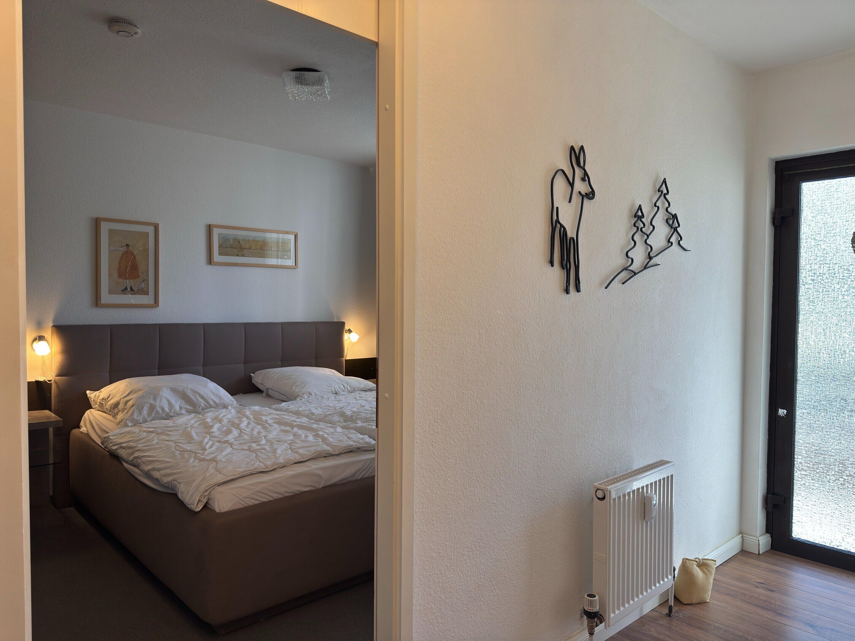 A bedroom with a bed, two wall-mounted lamps, a radiator, and two framed pictures on the wall.