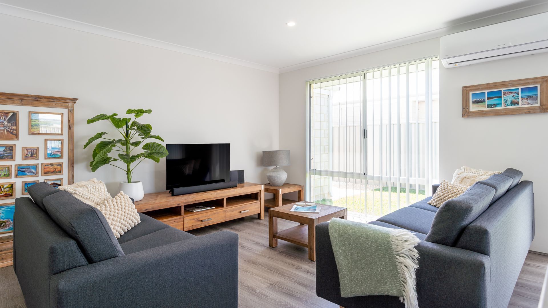 Un salon moderne avec deux canapés gris, un meuble TV en bois, une grande plante et une porte-fenêtre coulissante donnant sur un balcon.