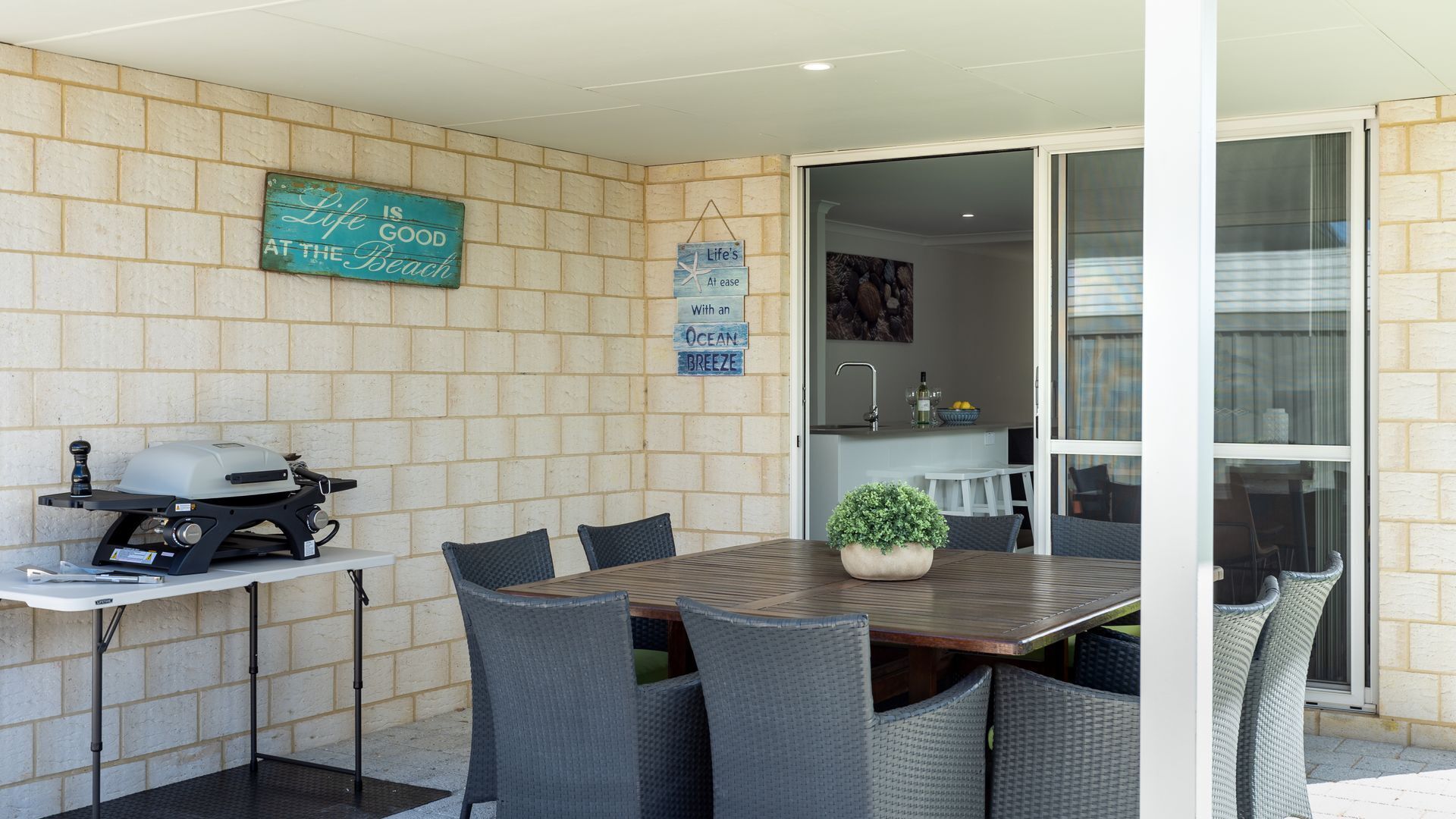An outdoor patio with a grill, dining table, and chairs, a kitchen area with a sink and counter, and a sliding glass door leading to an indoor space.