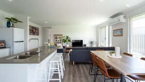 A modern kitchen with a central island, white bar stools, a dining table with chairs, a sofa, and a TV in the living area. - Whale of a Time - Vasse: Your Perfect Retreat for Comfort and Nature (Vasse)