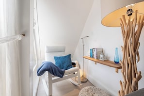 Um quarto com uma cadeira branca, um travesseiro azul, um abajur, uma prateleira de madeira com livros e um vaso, e uma estrutura decorativa de madeira.