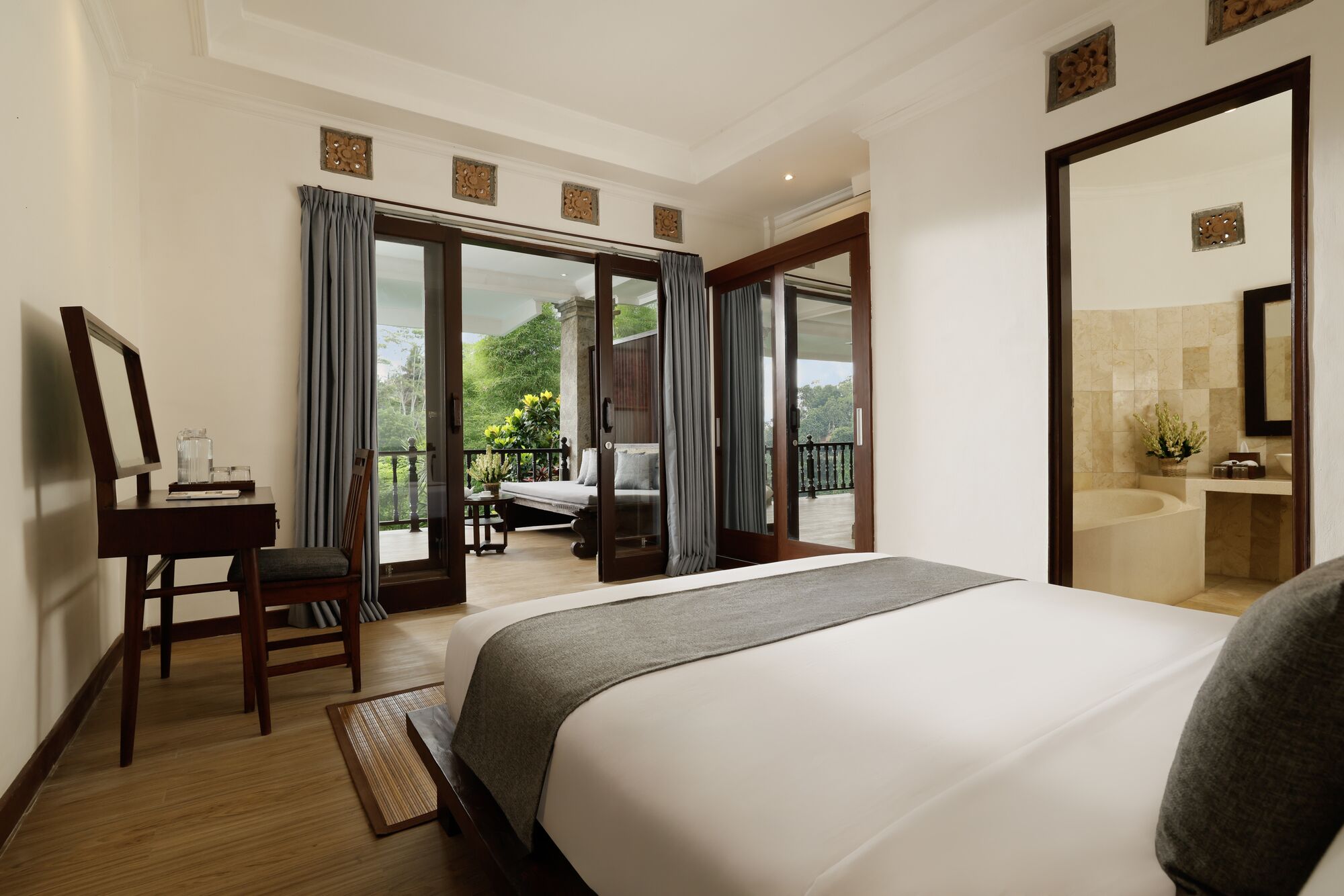 Une chambre d’hôtel avec un grand lit, un bureau avec une chaise, un balcon donnant sur un espace verdoyant, et une salle de bain avec une baignoire et un lavabo.
