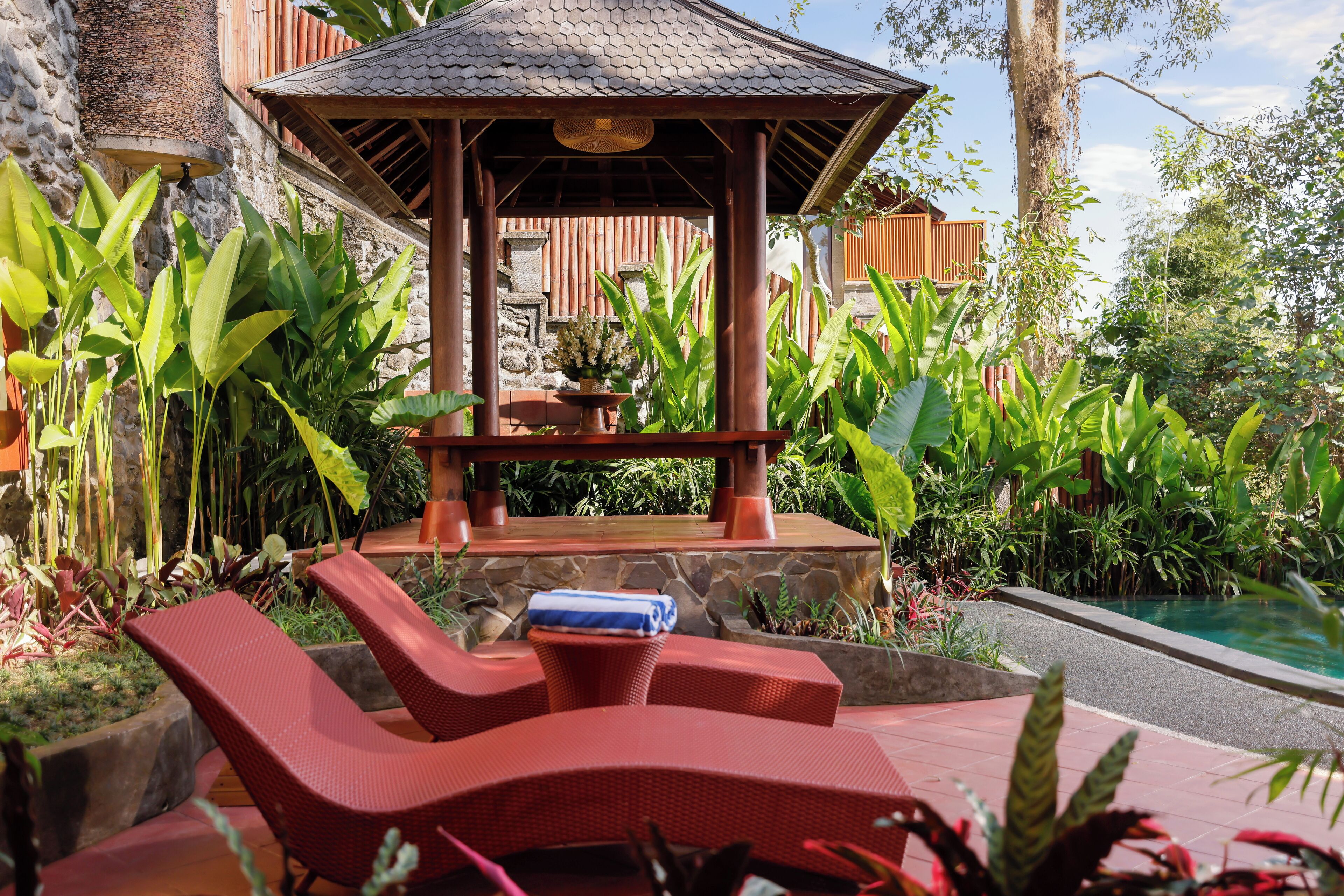 Ein Pavillon mit Steinunterbau, umgeben von üppiger tropischer Vegetation und einem Pool.