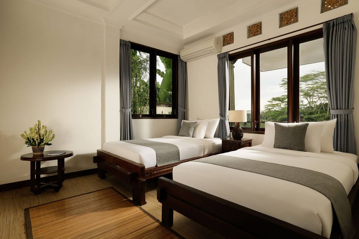 A hotel room with two beds, a bedside table with a lamp, a small table with a plant, and large windows with curtains.