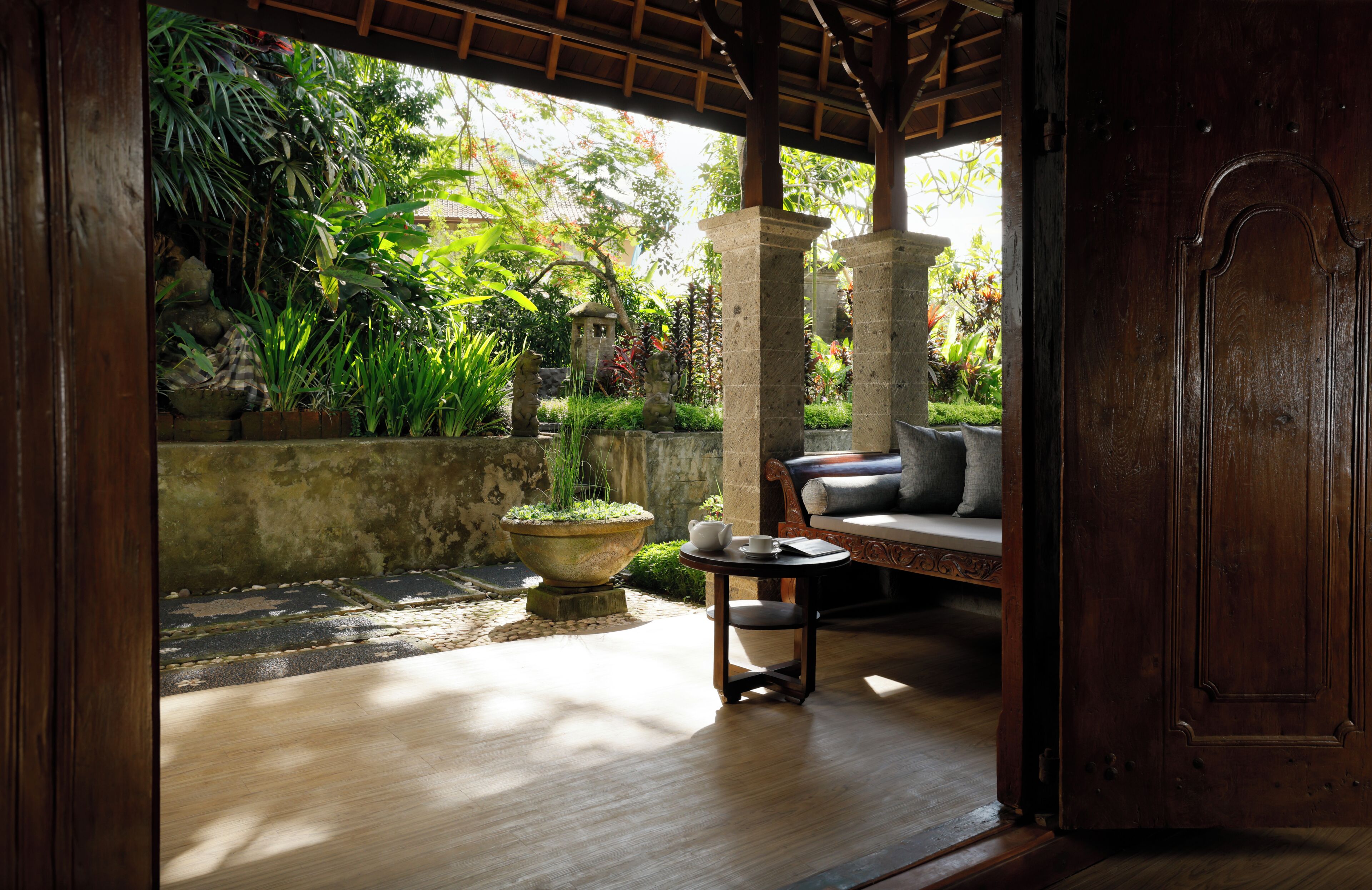 An open wooden door leading to a room with a stone bench, a small table with a cup and saucer, and a view of a lush garden.