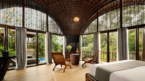 Quarto espaçoso com uma cama grande, duas cadeiras de madeira, uma mesa pequena e vista para vegetação exuberante através de amplas janelas.