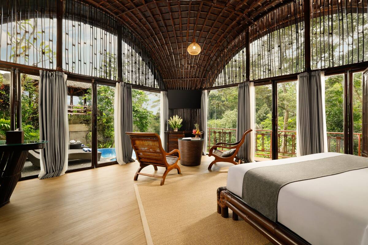 A spacious room with a large bed, two wooden chairs, a small table, and a view of lush greenery through large windows.