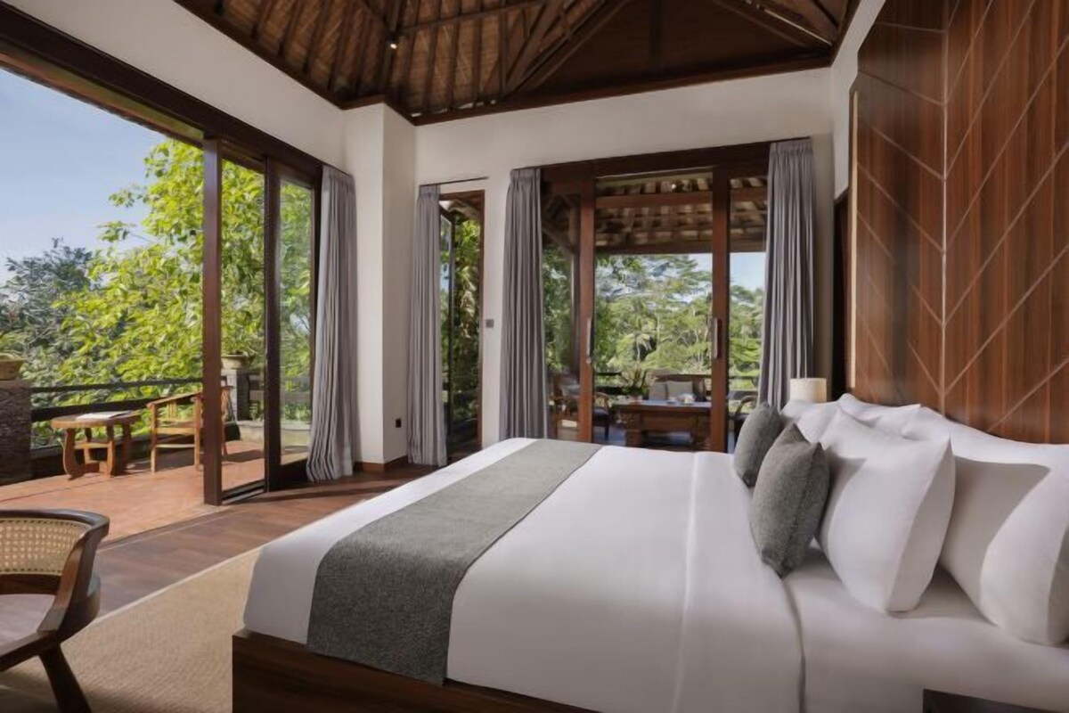 A spacious bedroom with a large bed, a balcony with a table and chairs, and a view of greenery.