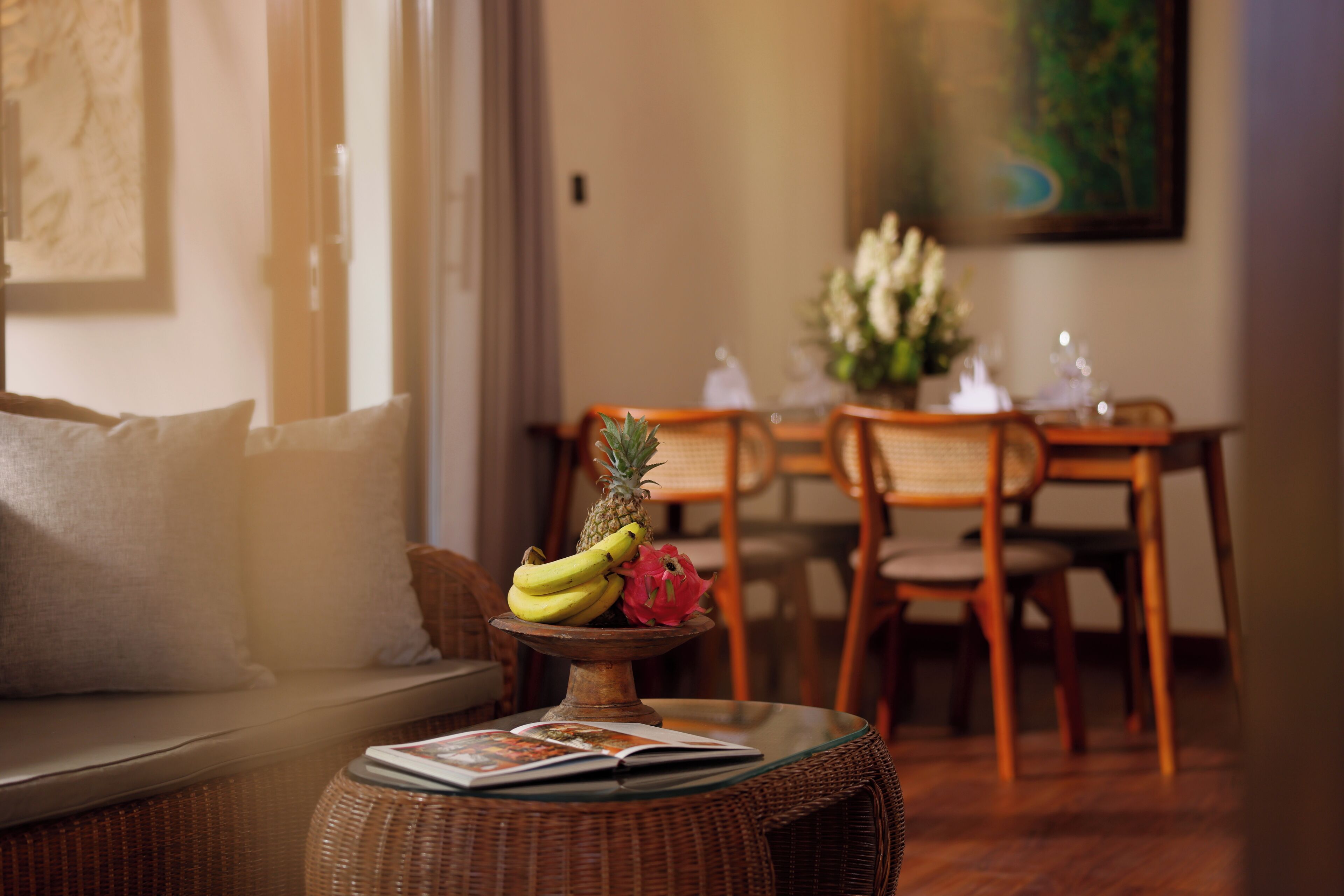 A dining area with a wicker table, a bowl of fruit, and a sofa with cushions.