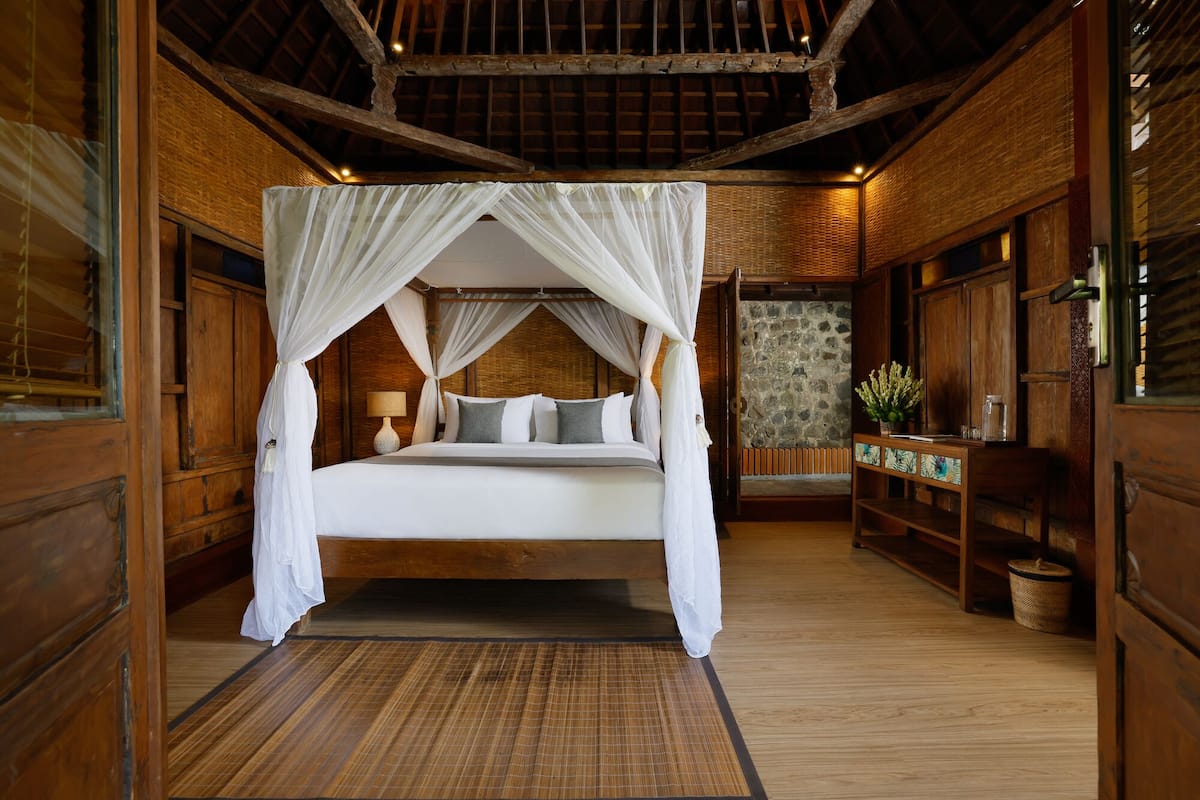 A wooden room with a bed under a canopy, a stone wall, and a wooden bench with a plant.