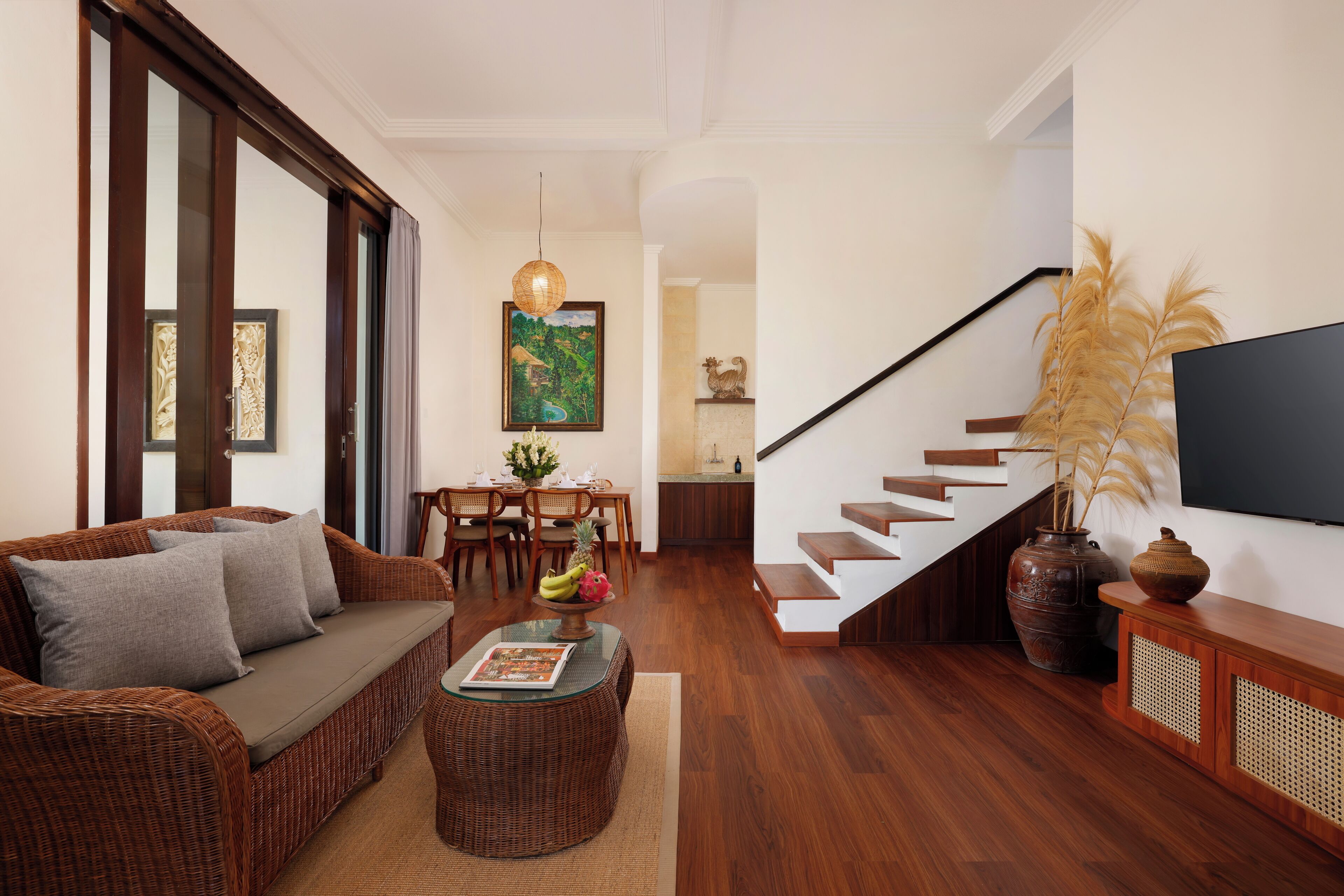 A modern living room with a wicker sofa, a dining table, and a staircase leading to an upper level.