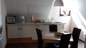 Una cocina pequeña con una mesa y sillas, un refrigerador blanco, una cafetera y una ventana con una planta.