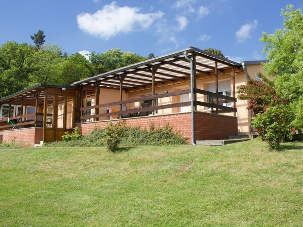 Un edificio de una planta con fachada de ladrillo, una terraza de madera y una pérgola con techo de metal.