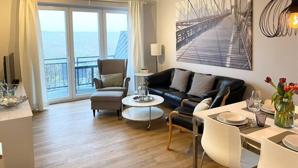 A modern living room with a large window, a black leather sofa, a checkered armchair, a small round table, and a dining area with a white table set. - Ferienwohnung, St. Peter - Ording (St. Peter-Ording)