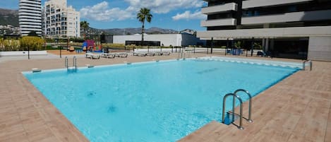 A rectangular swimming pool with a tiled deck, surrounded by modern buildings and a palm tree.