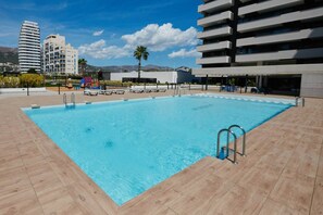 A rectangular swimming pool with a tiled deck, surrounded by modern buildings and a palm tree. - Alexia Apartment (Calpe)