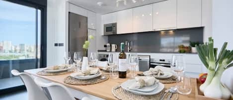 Un comedor moderno con una mesa de madera lista para una comida, sillas blancas y vistas a la ciudad a través de ventanas amplias.
