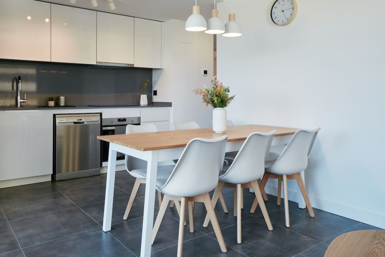 A modern kitchen with white cabinets, a stainless steel dishwasher, and a wooden dining table with four chairs.