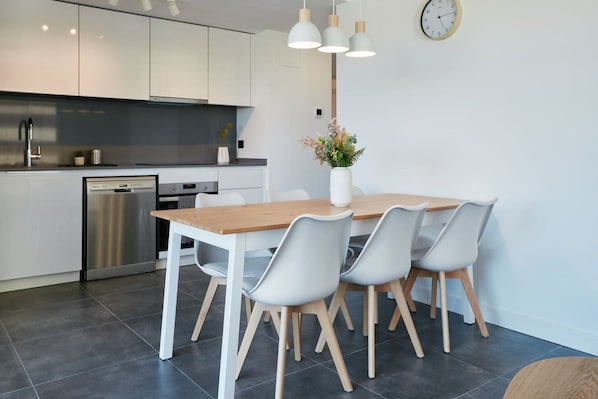 A modern kitchen with white cabinets, a stainless steel dishwasher, and a wooden dining table with four chairs.