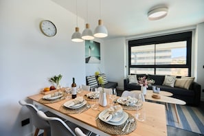 Un coin repas avec une table en bois dressée pour quatre personnes, une horloge au mur et un canapé à l’arrière-plan.
