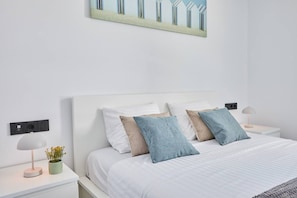 Une chambre à coucher moderne avec un lit blanc, deux tables de chevet, une prise électrique fixée au mur, une petite plante en pot et un tableau au-dessus du lit.
