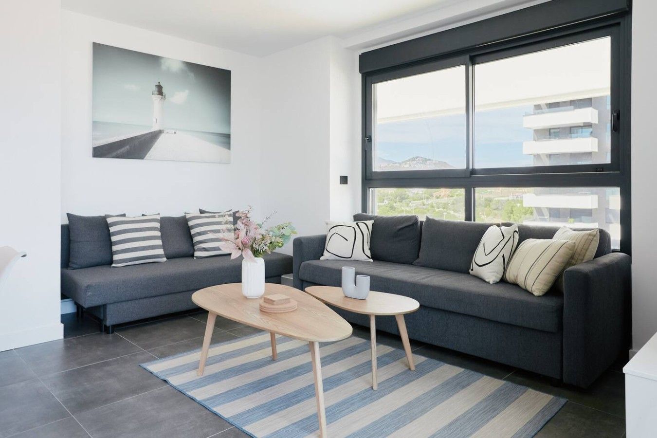 A modern living room with two grey sofas, a wooden coffee table, and a large window with a view of buildings and hills.