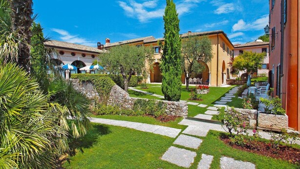 A courtyard with a stone pathway, palm trees, and a building with a tiled roof. - Residence La Filanda in Costermano (Costermano sul Garda)