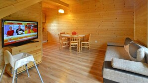 L’intérieur d’un chalet en bois comprend une télévision, une table à manger, des chaises, un canapé et un plafond haut avec des poutres apparentes.