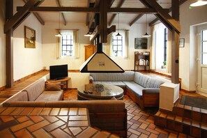 Una sala de estar amplia con un sofá grande, una mesa de centro y un televisor. La habitación tiene vigas de madera en el techo y ventanas grandes con cortinas.